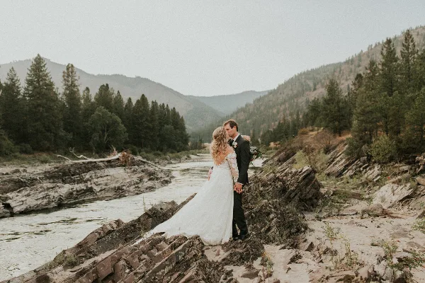 A bride and groom embracing on a rocky ledge overlooking a river and forested mountains.