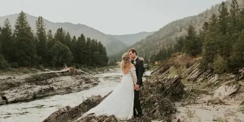 A bride and groom embracing on a rocky ledge overlooking a river and forested mountains.