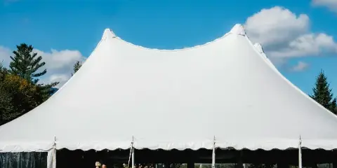 A large white peaked event tent set up outdoors against a blue sky with trees in the background.
