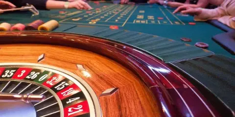 Close-up of a casino roulette wheel and green felt gaming table with chips and players' hands.