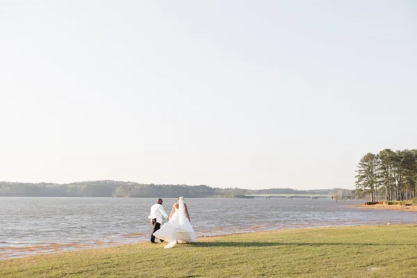 A bride and groom walking hand in hand along a grassy shoreline beside a lake with a bridge and trees in the distance.