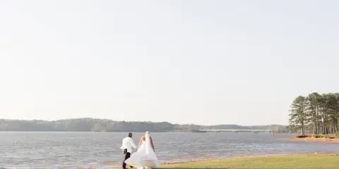 A bride and groom walking hand in hand along a grassy shoreline beside a lake with a bridge and trees in the distance.