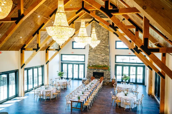 Interior view of a wedding venue featuring high wooden beams, crystal chandeliers, a stone fireplace, and set dining tables.