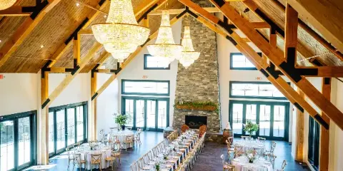 Interior view of a wedding venue featuring high wooden beams, crystal chandeliers, a stone fireplace, and set dining tables.