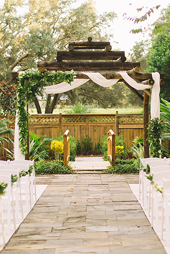 A stone pathway leading to a rustic wooden ceremony pergola decorated with greenery and white fabric, flanked by white chairs in a garden setting.