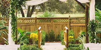 A stone pathway leading to a rustic wooden ceremony pergola decorated with greenery and white fabric, flanked by white chairs in a garden setting.
