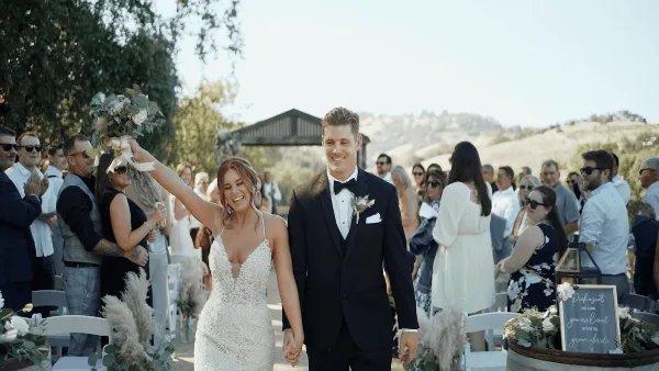 A bride and groom walking down the aisle at an outdoor wedding ceremony, smiling and holding hands while guests applaud.