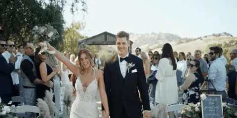 A bride and groom walking down the aisle at an outdoor wedding ceremony, smiling and holding hands while guests applaud.