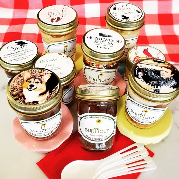 An assortment of Sunflour Baking Company cake jars with custom lids arranged on a red and white checkered tablecloth.