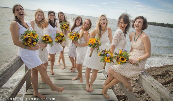 A group of bridesmaids in white dresses holding colorful sunflower and garden flower bouquets on a wooden dock.