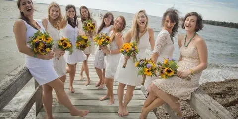 A group of bridesmaids in white dresses holding colorful sunflower and garden flower bouquets on a wooden dock.