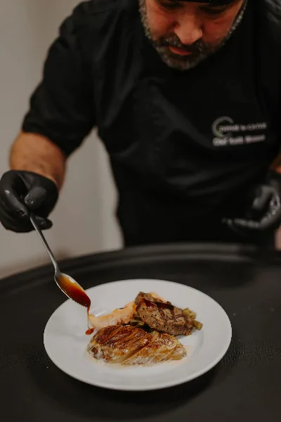 A chef in a black apron with the Thyme to Cater logo is carefully spooning sauce over a plated meal.