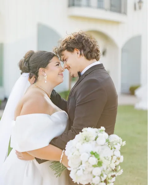 A bride and groom embracing and looking into each other's eyes outdoors, with the bride holding a white bouquet.