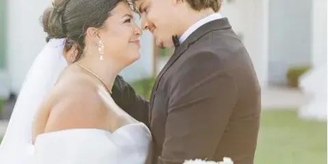 A bride and groom embracing and looking into each other's eyes outdoors, with the bride holding a white bouquet.