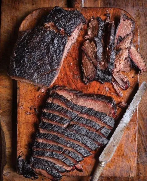 A large, wood-smoked roast with a dark crust, partially sliced on a rustic wooden cutting board.