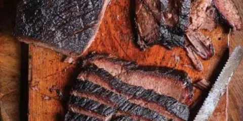 A large, wood-smoked roast with a dark crust, partially sliced on a rustic wooden cutting board.