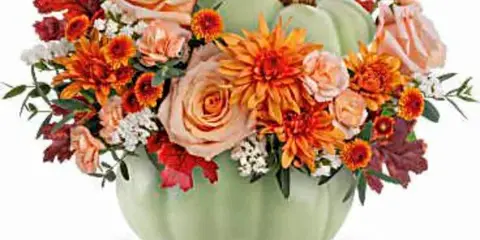 A floral arrangement featuring peach roses, orange daisies, and red maple leaves arranged in a green pumpkin-shaped vase.