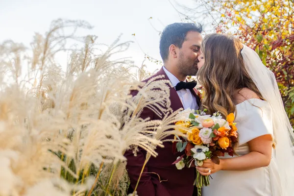 A bride and groom sharing a kiss outdoors surrounded by tall ornamental grasses and autumn foliage.
