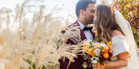 A bride and groom sharing a kiss outdoors surrounded by tall ornamental grasses and autumn foliage.