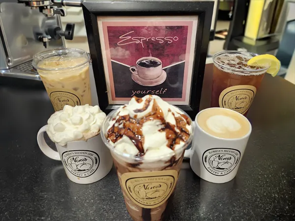 An assortment of iced and hot specialty coffee drinks, including lattes and frappes, served in branded cups and mugs on a cafe counter.