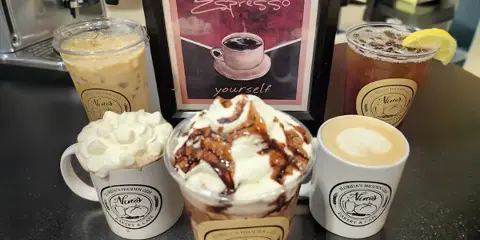 An assortment of iced and hot specialty coffee drinks, including lattes and frappes, served in branded cups and mugs on a cafe counter.