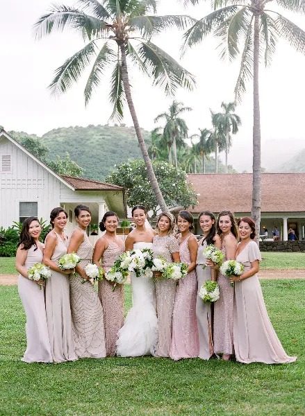 A bride and her bridesmaids posing for a group photo on a lawn with palm trees in the background.