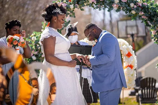 A bride and groom exchanging rings during an outdoor wedding ceremony under a floral arch.