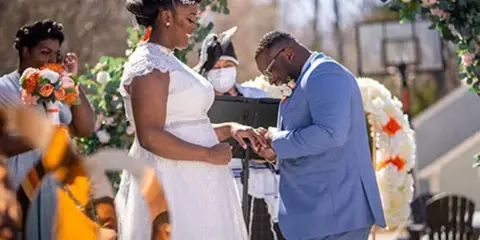 A bride and groom exchanging rings during an outdoor wedding ceremony under a floral arch.