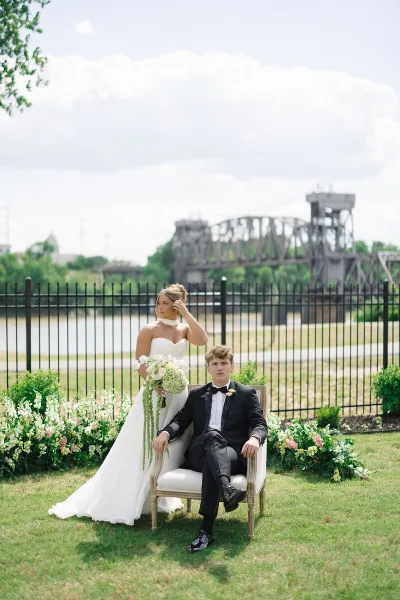 A bride and groom posing outdoors on a lawn with a river and bridge in the background.