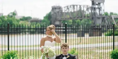 A bride and groom posing outdoors on a lawn with a river and bridge in the background.