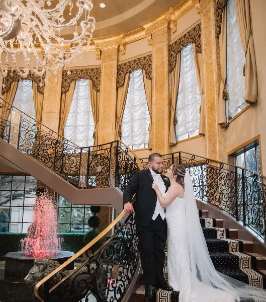 A bride and groom standing on a staircase in a luxurious ballroom with a red-lit indoor fountain.