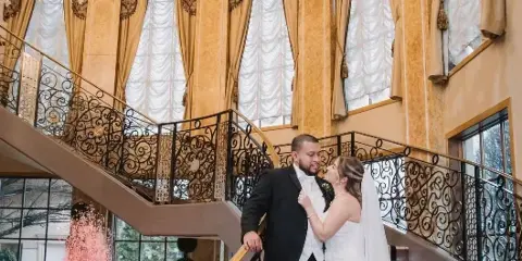 A bride and groom standing on a staircase in a luxurious ballroom with a red-lit indoor fountain.