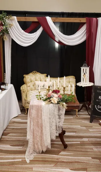 A wedding event setup featuring a cream-colored Victorian couch, a table with lace linens and candles, and a wooden backdrop with draped white and burgundy fabric.