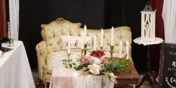 A wedding event setup featuring a cream-colored Victorian couch, a table with lace linens and candles, and a wooden backdrop with draped white and burgundy fabric.