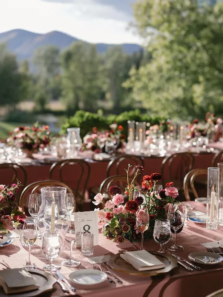 A close-up view of a wedding reception table featuring a lush floral arrangement with red and pink blooms, glassware, and a table number card, set against a mountain landscape.
