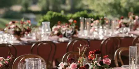 A close-up view of a wedding reception table featuring a lush floral arrangement with red and pink blooms, glassware, and a table number card, set against a mountain landscape.