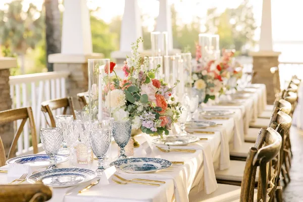 A long event table set with white linens, blue and white plates, gold flatware, and glassware, accompanied by floral centerpieces and wooden chairs.