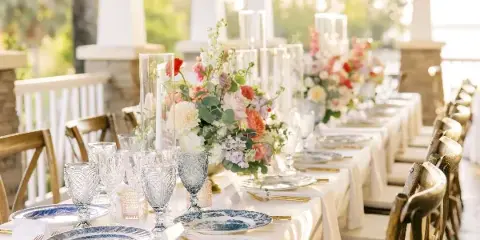 A long event table set with white linens, blue and white plates, gold flatware, and glassware, accompanied by floral centerpieces and wooden chairs.