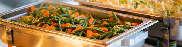 A close-up of a catering buffet featuring a stainless steel chafing dish filled with sautéed green beans and carrots, with another dish visible in the background.