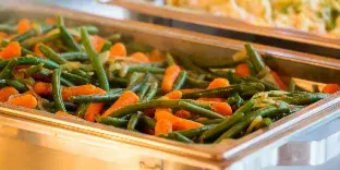 A close-up of a catering buffet featuring a stainless steel chafing dish filled with sautéed green beans and carrots, with another dish visible in the background.