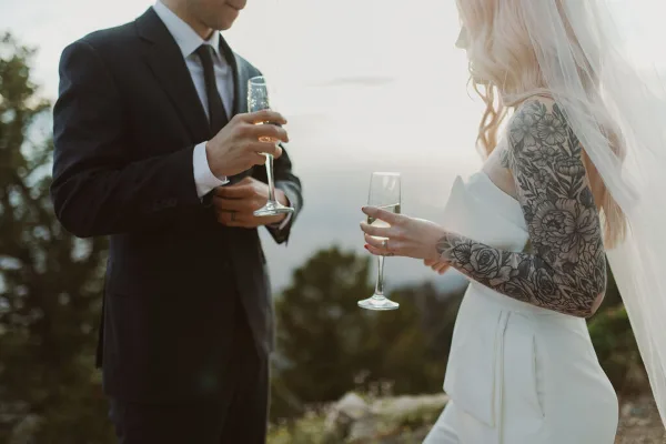 A bride and groom in wedding attire holding champagne flutes outdoors against a scenic background.