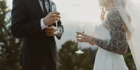 A bride and groom in wedding attire holding champagne flutes outdoors against a scenic background.