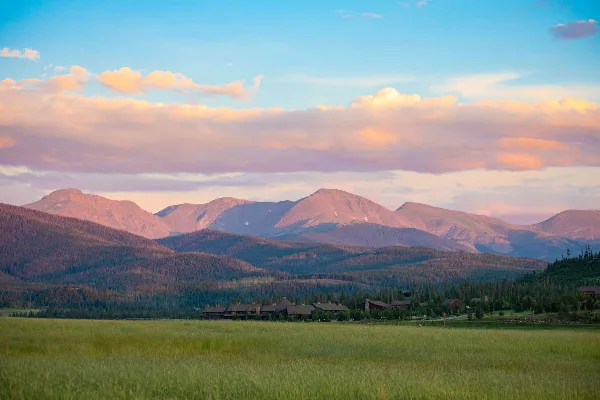 Wide landscape view of the Devil's Thumb Ranch resort buildings in a green meadow with forested hills and mountains in the background at sunset.