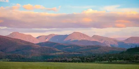 Wide landscape view of the Devil's Thumb Ranch resort buildings in a green meadow with forested hills and mountains in the background at sunset.