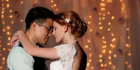 A bride and groom embracing in front of a backdrop of warm string lights.