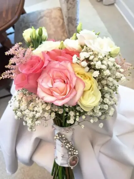 A hand-tied bridal bouquet with pink and yellow roses, white lisianthus, and baby's breath, wrapped in a white ribbon with a jeweled brooch.