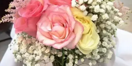 A hand-tied bridal bouquet with pink and yellow roses, white lisianthus, and baby's breath, wrapped in a white ribbon with a jeweled brooch.