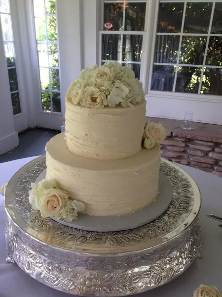 A two-tier white wedding cake decorated with cream roses and hydrangeas, displayed on a silver pedestal stand.