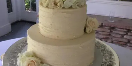 A two-tier white wedding cake decorated with cream roses and hydrangeas, displayed on a silver pedestal stand.