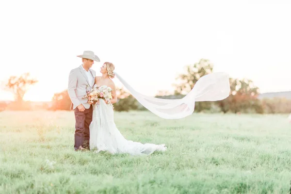 A bride and groom standing in a grassy field at sunset, with the bride's veil blowing in the wind.
